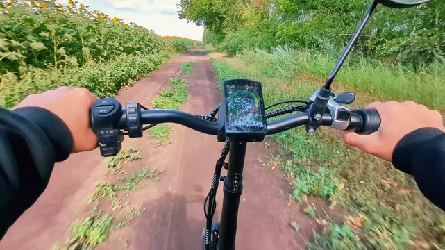 A man rides an electric bicycle from a first person perspective along a dirt road in the middle of a sunflower field, with a row of birch trees growing on the right side. POV