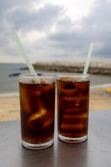 glass of iced cola sits on a table by the sea, with a cloudy sky backdrop. The drink's bubbles and condensation are visible