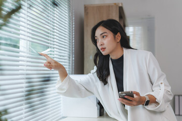 Businesswoman opening venetian blinds and holding smartphone in office