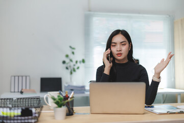Stressed asian businesswoman talking on phone and working on laptop in office