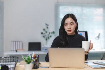Focused asian businesswoman drinking coffee and working on laptop in modern office