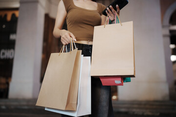 Stylish young woman walking outdoors at night, carrying shopping bags with joy, surrounded by city...