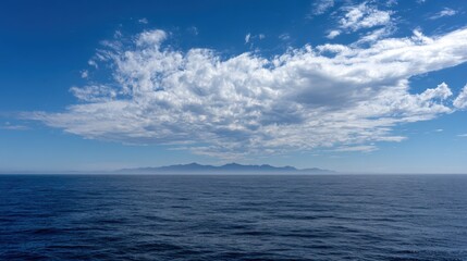 Obraz premium Bright clouds drift lazily across a vast blue sky above tranquil ocean waters. In the distance, a rugged island silhouette emerges against the horizon, hinting at natural beauty.