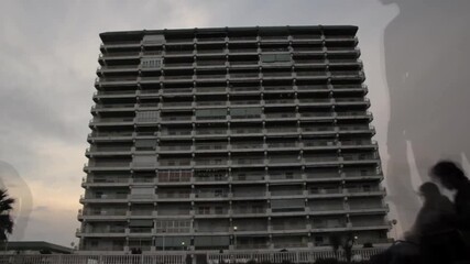 Ghostlike figures drift past a massive apartment block at dusk, blending memory and reality—evoking nostalgia, presence, and the layers of urban life.