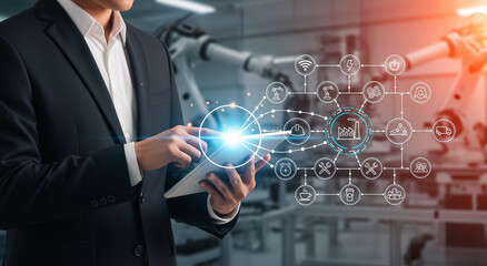 Businessman using a tablet to control a smart factory system with robotic arms in the background, symbolizing industry 4.0, automation, and digital manufacturing