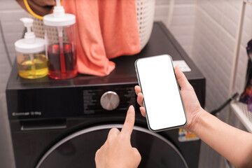 Hand holding and pointing at blank smartphone screen in laundry room near washing machine and detergent bottles