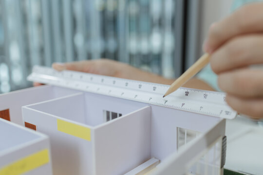 Asian male architect sitting at a desk, measuring a modern house model with triangular ruler and pencil. Office setup includes design tools, blueprints, and safety gear, suggesting architectural work.