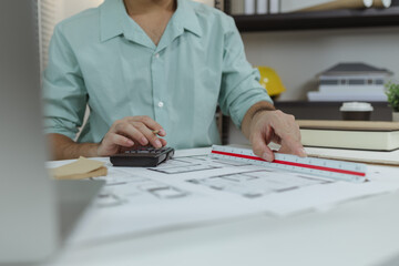 An architect working on house plans at a white desk with blueprints, a calculator, and a triangular scale ruler, focusing on drawing and measuring details in a clean, modern workspace.