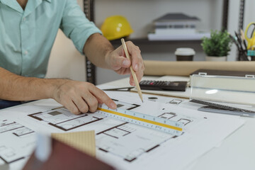 An architect using a pencil and a scale ruler to measure house floor plans. Various design tools and model pieces are on the desk, indicating active architectural planning process.