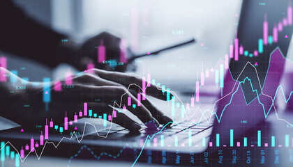 Hands typing on laptop keyboard with glowing stock market charts and financial graphs overlayed in digital interface concept on office desk background.
