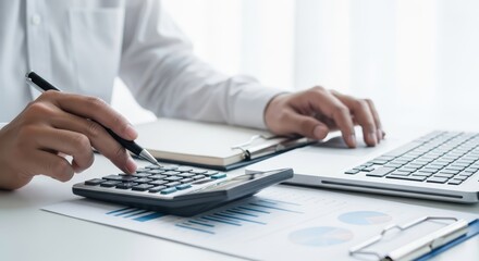 A person's hands using a calculator and typing on a laptop keyboard with financial reports and a notebook on a white desk