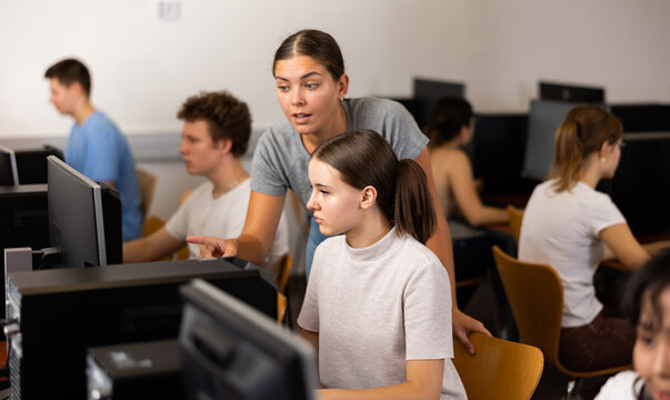 Young experienced female teacher helping girl pupil working with computer in classroom of college