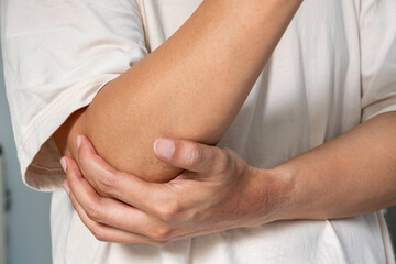 A person grips their elbow, indicating localized pain or strain. The light indoor setting suggests a minor injury, possibly from repetitive use or sudden motion. Healthcare and medical concept.