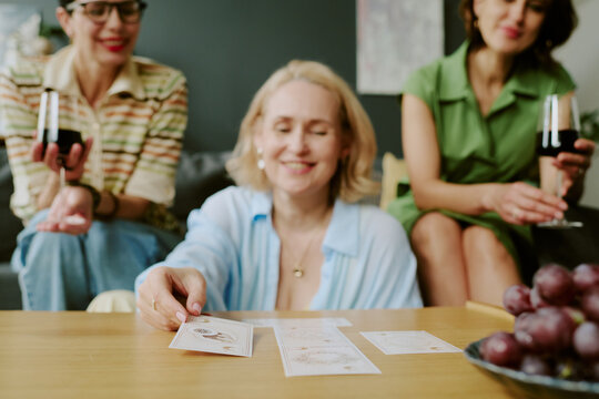 Three Caucasian women sitting together, smiling and holding wine glasses, middle aged woman in foreground placing tarot cards on table, group engaging in tarot reading activity