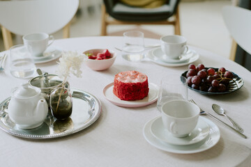 Round table set with porcelain teapot, cups, saucers, glassware, small red cake on plate, bowl of watermelon cubes, plate of grapes, floral arrangement in glass vase