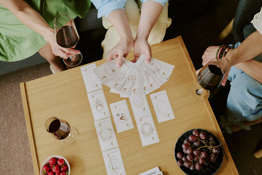 Three young adult Caucasian women sitting around table playing tarot card game, holding wine glasses, arranging cards, sharing relaxed moment, fruit bowls visible on tabletop