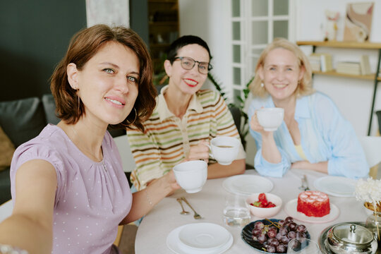Portrait of three middle aged Caucasian women smiling and holding cups while sitting at table with desserts and fruit, one woman taking selfie, all looking at camera