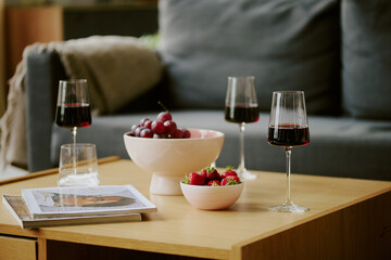 Closeup showing three glasses of red wine, bowl of grapes, bowl of strawberries, two magazines and two water glasses on wooden coffee table in living room setting