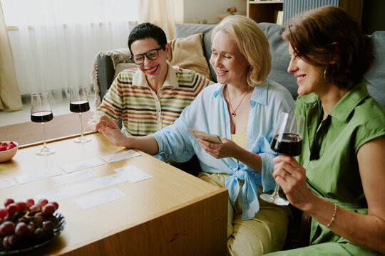 Three middle aged Caucasian women sitting together playing card game at table, smiling and holding glasses of red wine, enjoying leisure time in living room setting