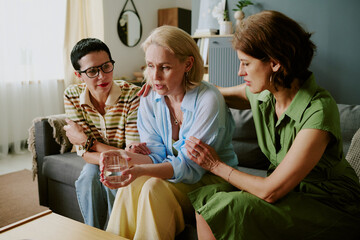 Middle aged Caucasian woman sitting on sofa holding glass of water looking distressed while two middle aged close friends comforting her with supportive gestures