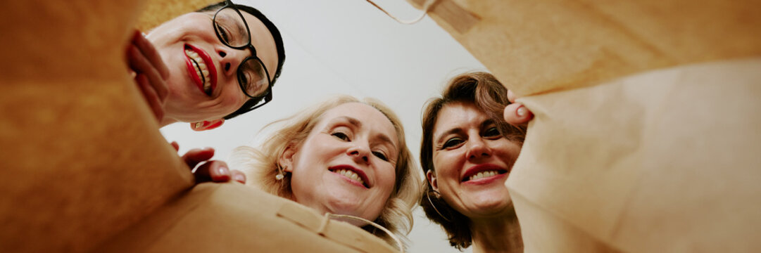 Three middle aged Caucasian women smiling and looking down into shopping bag, faces close together, showing joyful expressions, photographed from inside bag perspective