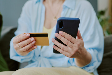 Unrecognizable woman holding smartphone in one hand and credit card in other hand, making online payment or shopping transaction, sitting indoors