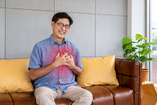 Asian elderly man suffering from chest pain, possible heart attack, while sitting on a leather sofa at home