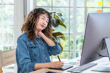 Asian female graphic designer with neck pain at desk using drawing tablet and computer, office...