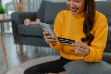 Asian woman holding a smartphone and credit card while making an online purchase at home. She sits...