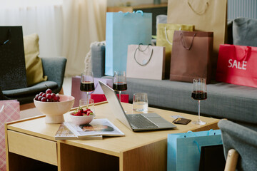 Shopping bags surrounding wooden table with open laptop, glasses of red wine, bowl of grapes, strawberries, magazine and smartphone, suggesting recent shopping activity at home