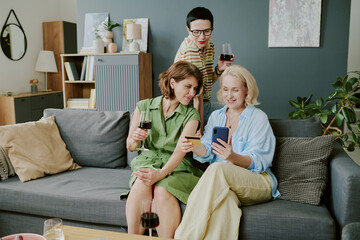 Middle aged Caucasian girlfriends sitting on sofa in cozy living room, looking at smartphone screen and drinking wine