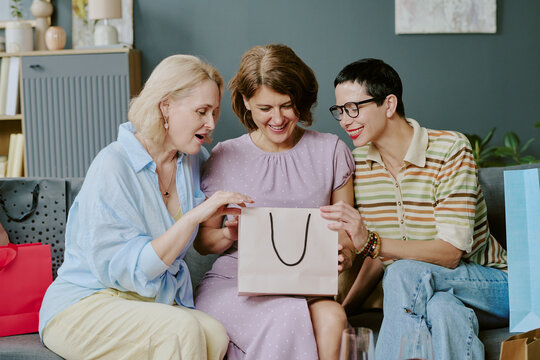 Three middle aged Caucasian women sitting together on sofa smiling and opening shopping bag, engaging in friendly conversation during leisure time