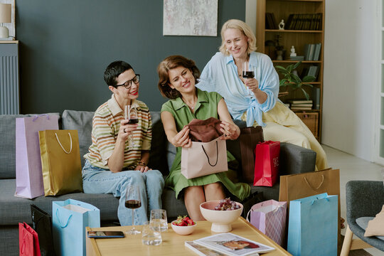 Three middle aged Caucasian women sitting on sofa smiling and holding wine glasses, one woman opening shopping bag surrounded by multiple colorful shopping bags and snacks on table