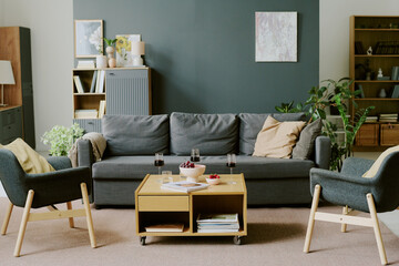 Modern living room interior featuring gray sofa, two armchairs, wooden coffee table with bowl of fruit, glasses of red wine, books and green plants arranged in background