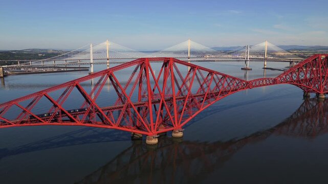 Aerial video of a train travelling over the Forth Bridge in Scotland, with Forth Road Bridge and Queensferry Crossing on the background. 