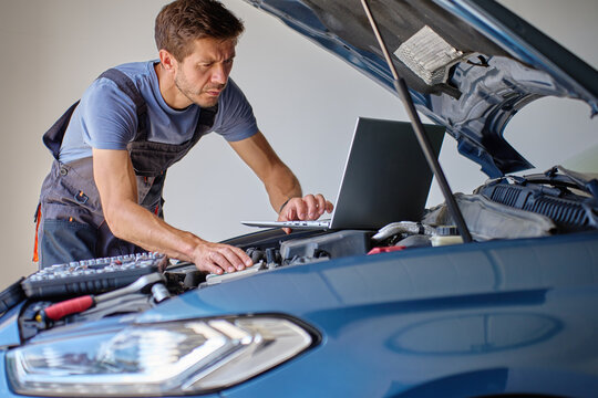 Auto mechanic using laptop for diagnostics vehicle engine in auto repair shop. Man in work overalls standing near car with open hood in garage. Vehicle maintenance and diagnostics concept