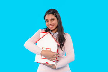 Young woman smiling while holding a miniature house against a bright blue background, symbolizing...