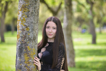 Latin woman, young and beautiful brunette dressed in black is clinging and hiding behind a tree trunk in the forest. The woman looks shyly and sensually. Beauty and fashion concept.