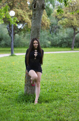 Latin woman, young and beautiful brunette dressed in black leans back against the trunk of a tree in the forest. The woman looks shyly and sensually. Beauty and fashion concept.