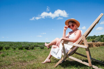 Young beautiful sexy woman in straw hat and sunglasses sitting on a sun lounger on a meadow on a sunny day against the sky. Rest. Vacation. Tourism. Background.