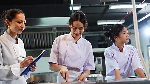 Female chef teacher teaches two students to cook in class. A cooking test is given to the teenage girls to cook spaghetti. The teacher holds a file to give scores in the cooking class.