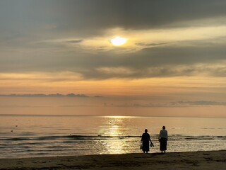 Fototapeta premium Two people walking along a calm beach at sunset under a dramatic sky, with golden sunlight reflecting on the sea and horizon.
