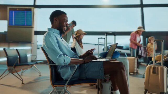 A man sits in an airport, using a laptop and a credit card, other travelers are in the background.