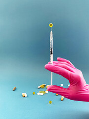 Syringe with yellow drop held by hand in pink medical glove, scattered pills and capsules on blue background, concept of medicine and healthcare