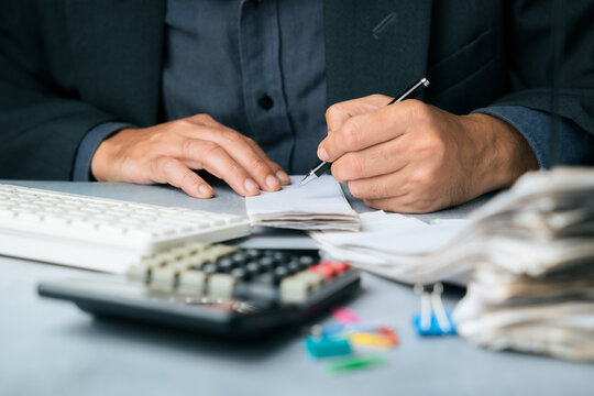 Businessman writing notes on a bill document at a desk with calculator, keyboard, and paper stacks, representing financial planning, accounting, or expense tracking tasks - Powered by Adobe