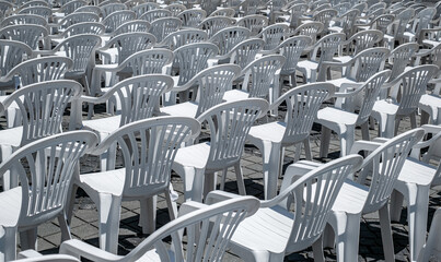 Obraz premium Photograph of an outdoor seating arrangement featuring numerous white plastic chairs and tables arranged in rows on a sunlit, cobblestone pavement