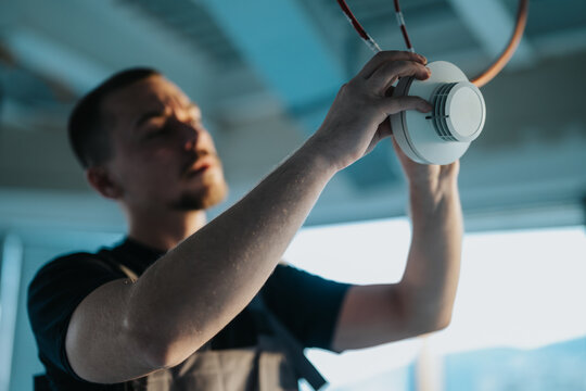 A professional technician is carefully installing a smoke detector on a ceiling, showcasing expertise and focus in fire safety and security systems in a workplace environment.