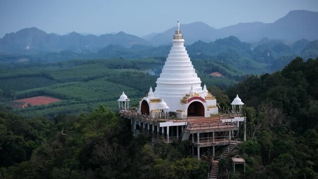 Drone footage of a white Buddhist temple atop a forested mountain in Surat Thani Province, Thailand. Midday light enhances the serene, spiritual landscape.