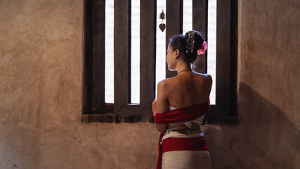 A beautiful, young Asian woman wearing a traditional Thai dress, Lanna style, stands beside an old...