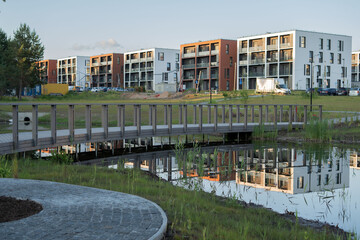 Fototapeta premium modern apartments with diverse facades reflected in a serene pond, connected by a brown wooden bridge in an urban park setting during clear daylight, surrounded by greenery, fresh, open, community